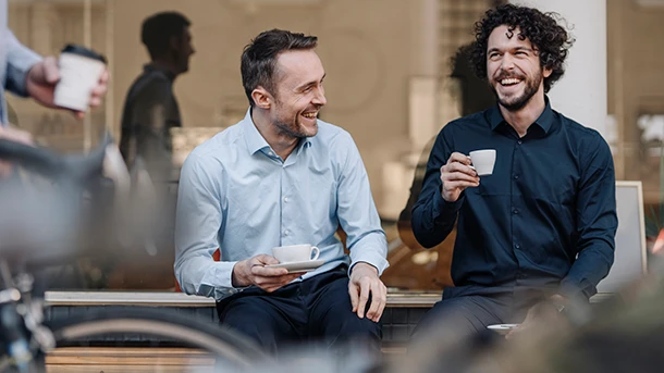 two men sitting on a bench and holding cups of coffee